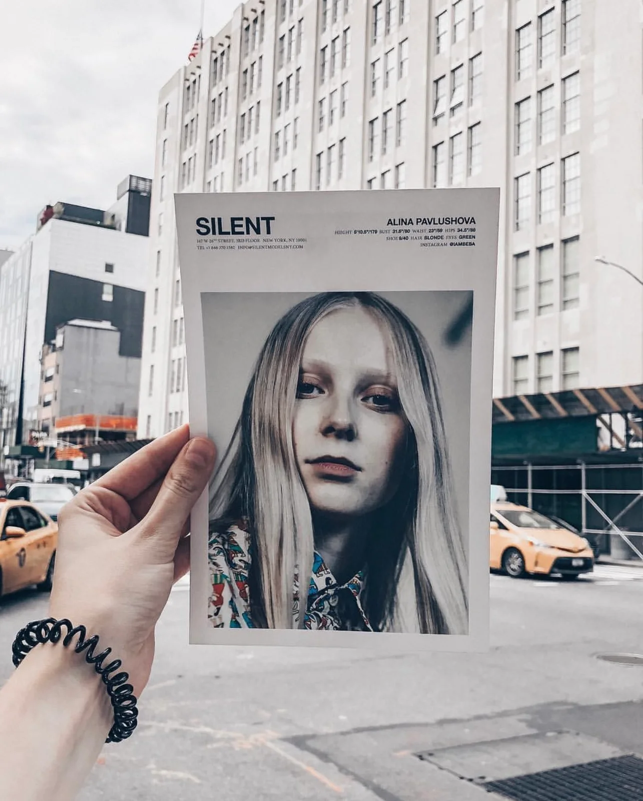 A hand holding Alina's SILENT model comp card on a New York street with yellow taxis and Manhattan buildings