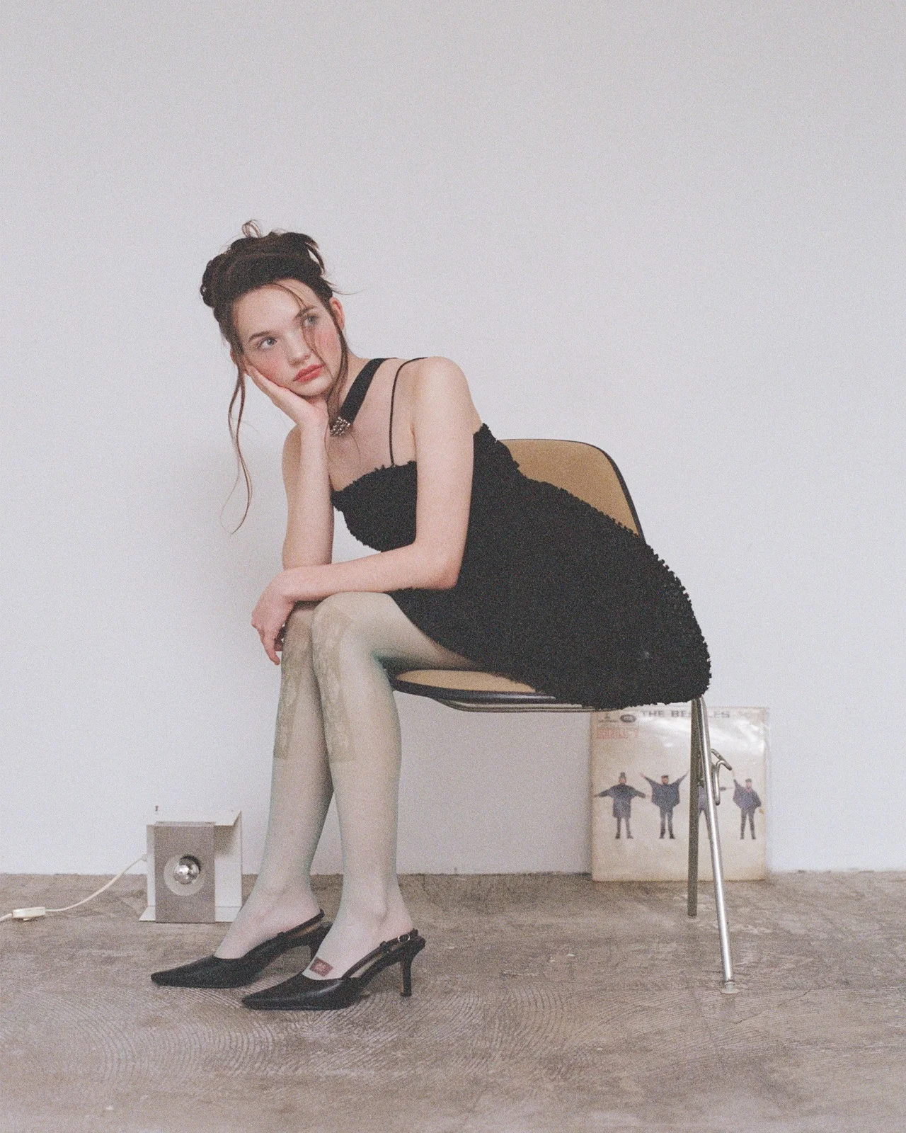 Ellie seated on a chair in a black dress and heels, resting her cheek on her hand