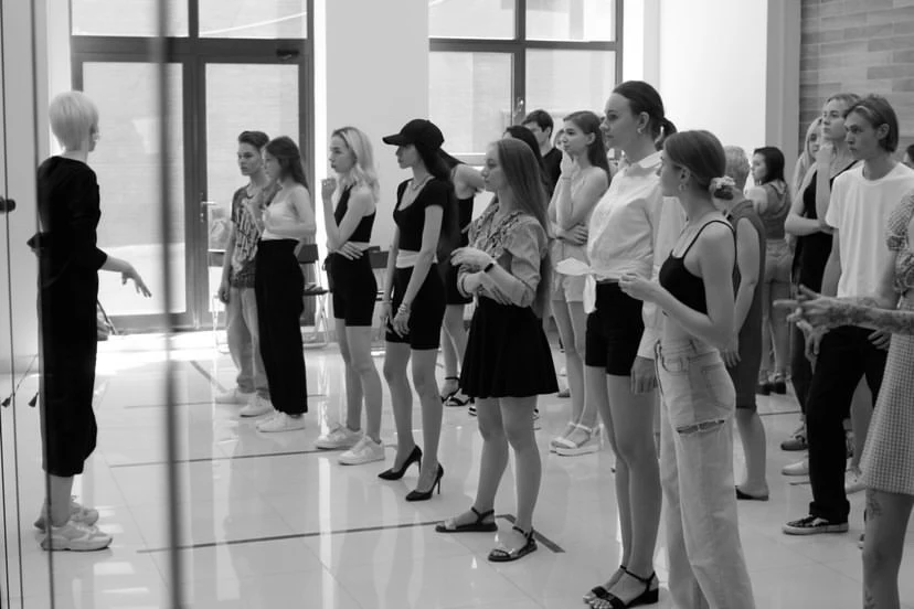 A modelling coach addressing a line of young models at a posing workshop, in black and white